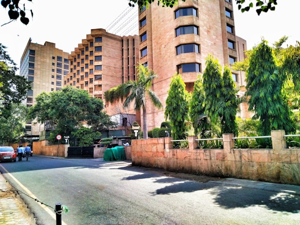 A modern cityscape featuring a large building with unique architectural design elements including geometric patterns and large windows. The surrounding area is landscaped with various trees and shrubs, providing a green contrast to the beige and brown tones of the building. There are a few people walking along the empty street alongside the building.