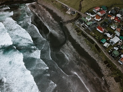 An aerial view of a coastal area with rough waves crashing onto a rocky shoreline. To the right, there is a small village with houses featuring colorful roofs. The landscape includes a combination of grass, rocks, and concrete pathways.
