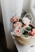 A woven basket filled with soft pink artificial flowers and green leaves is placed on a white surface near a sheer curtain. Delicate white baby's breath flowers complement the arrangement, adding a touch of elegance to the floral display.