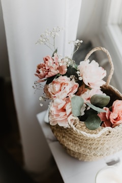 A woven basket filled with soft pink artificial flowers and green leaves is placed on a white surface near a sheer curtain. Delicate white baby's breath flowers complement the arrangement, adding a touch of elegance to the floral display.