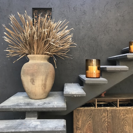 A rustic, textured wall serves as the background for a decorative arrangement. An earthy, clay vase filled with dried grasses sits on a stepped stone ledge. Beside it, a series of candles in brown glass holders are placed on the stone steps, adding a warm, natural ambiance.