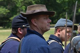 Several men wearing historical military uniforms stand outdoors. They are dressed in dark blue clothing with hats, and carry long bayonets. The background is filled with green foliage, indicating a natural setting.