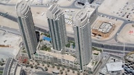 Aerial view of three high-rise buildings surrounded by desert landscape and roads. The buildings are tall with modern architecture, featuring balconies and large windows. Below, there are landscaped areas with palm trees and a swimming pool visible within the complex.