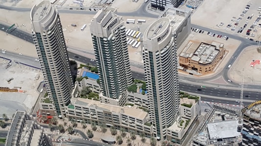 Aerial view of three high-rise buildings surrounded by desert landscape and roads. The buildings are tall with modern architecture, featuring balconies and large windows. Below, there are landscaped areas with palm trees and a swimming pool visible within the complex.