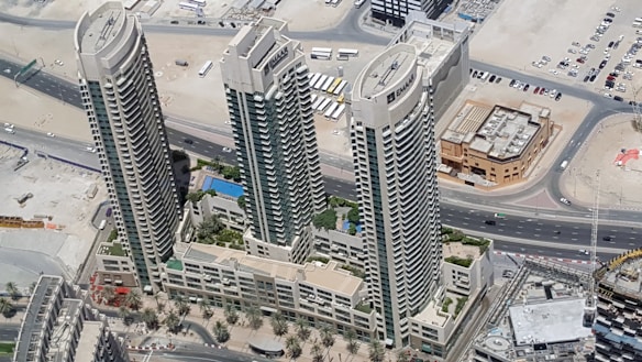 Aerial view of three high-rise buildings surrounded by desert landscape and roads. The buildings are tall with modern architecture, featuring balconies and large windows. Below, there are landscaped areas with palm trees and a swimming pool visible within the complex.