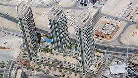 Aerial view of three high-rise buildings surrounded by desert landscape and roads. The buildings are tall with modern architecture, featuring balconies and large windows. Below, there are landscaped areas with palm trees and a swimming pool visible within the complex.
