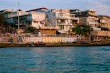 A coastal scene featuring several modern buildings lined along the shore with balconies and large windows. A colorful wall mural with the word 'Bondi' is visible near the water's edge. People walk along the shore, enjoying the scenic view. The ocean in the foreground reflects the evening sunlight.