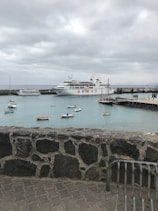 A harbor scene featuring a large passenger ferry with the name 'ARMAS' docked at a pier. Several small boats are floating on calm turquoise waters. The sky is overcast with a thick layer of gray clouds. A stone wall and part of a metal bench are visible in the foreground.
