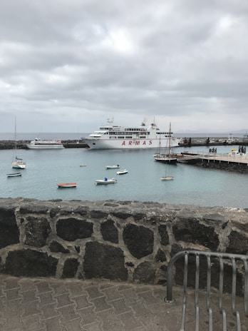 A harbor scene featuring a large passenger ferry with the name 'ARMAS' docked at a pier. Several small boats are floating on calm turquoise waters. The sky is overcast with a thick layer of gray clouds. A stone wall and part of a metal bench are visible in the foreground.