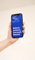 A hand holding a smartphone with a blue screen displaying the motivational text: 'Believe, Achieve, Succeed, Repeat.'