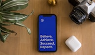 A smartphone with a bright blue screen displaying the motivational words 'Believe, Achieve, Succeed, Repeat' is placed on a wooden surface. Surrounding the phone are a potted plant with striped leaves, a vintage camera, and a wireless earbud case.