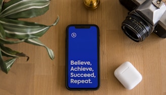 A smartphone with a bright blue screen displaying the motivational words 'Believe, Achieve, Succeed, Repeat' is placed on a wooden surface. Surrounding the phone are a potted plant with striped leaves, a vintage camera, and a wireless earbud case.
