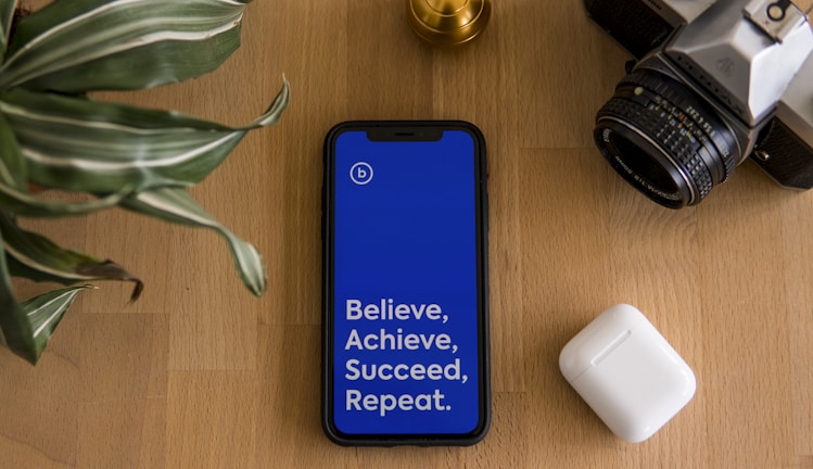A smartphone with a bright blue screen displaying the motivational words 'Believe, Achieve, Succeed, Repeat' is placed on a wooden surface. Surrounding the phone are a potted plant with striped leaves, a vintage camera, and a wireless earbud case.