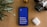 A smartphone with a bright blue screen displaying the motivational words 'Believe, Achieve, Succeed, Repeat' is placed on a wooden surface. Surrounding the phone are a potted plant with striped leaves, a vintage camera, and a wireless earbud case.