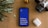 A smartphone with a bright blue screen displaying the motivational words 'Believe, Achieve, Succeed, Repeat' is placed on a wooden surface. Surrounding the phone are a potted plant with striped leaves, a vintage camera, and a wireless earbud case.