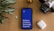 A smartphone with a bright blue screen displaying the motivational words 'Believe, Achieve, Succeed, Repeat' is placed on a wooden surface. Surrounding the phone are a potted plant with striped leaves, a vintage camera, and a wireless earbud case.