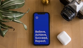 A smartphone with a bright blue screen displaying the motivational words 'Believe, Achieve, Succeed, Repeat' is placed on a wooden surface. Surrounding the phone are a potted plant with striped leaves, a vintage camera, and a wireless earbud case.