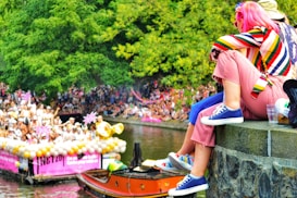 A vibrant outdoor scene of a festive canal parade with a diverse crowd in colorful attire. Several people are watching from a stone ledge, including an individual in a bright striped sweater and pink pants. A decorated boat with balloons and performers in white is passing by, while lush green trees provide a natural backdrop.