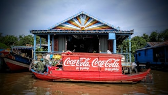 A colorful floating market stall on a canal features a prominent Coca-Cola logo on a red boat. The structure behind the boat has a vibrant, multi-colored triangular design on its roof. Several goods, including beverages and snacks, are displayed, with a person managing the stall. Lush green trees surround the area, contributing to the scenic atmosphere.