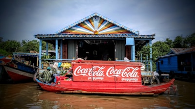 A colorful floating market stall on a canal features a prominent Coca-Cola logo on a red boat. The structure behind the boat has a vibrant, multi-colored triangular design on its roof. Several goods, including beverages and snacks, are displayed, with a person managing the stall. Lush green trees surround the area, contributing to the scenic atmosphere.