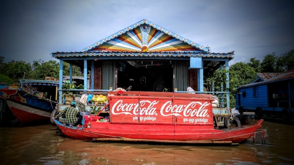 A colorful floating market stall on a canal features a prominent Coca-Cola logo on a red boat. The structure behind the boat has a vibrant, multi-colored triangular design on its roof. Several goods, including beverages and snacks, are displayed, with a person managing the stall. Lush green trees surround the area, contributing to the scenic atmosphere.
