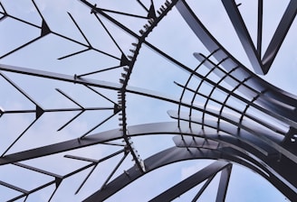 Intricate architectural structure composed of intersecting metal beams arranged in a radial pattern against a clear blue sky. The beams form a complex geometric design with varying angles and curves, creating a sense of depth and modernity.