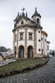 An ornate, historic church stands prominently with arched entrances and a cross atop its baroque architecture. The structure is set on a slightly elevated cobblestone path, surrounded by a lush, green landscape. The building features intricate stonework and multiple circular windows.
