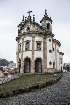 An ornate, historic church stands prominently with arched entrances and a cross atop its baroque architecture. The structure is set on a slightly elevated cobblestone path, surrounded by a lush, green landscape. The building features intricate stonework and multiple circular windows.