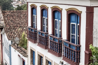 A colonial-style building with blue and white painted wooden components and arched windows. The exterior features intricate wooden railings and a rooftop covered in aged ceramic tiles. There's a street lamp visible, surrounded by lush greenery in the background.