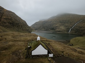 A small, white church with a grass roof is situated in a vast, open landscape characterized by large, rugged hills and a foggy sky. A waterfall cascades down one of the hills into a calm body of water, contributing to the serene and remote atmosphere. The terrain surrounding the church is grassy and uneven, with a few scattered rocks and pathways.