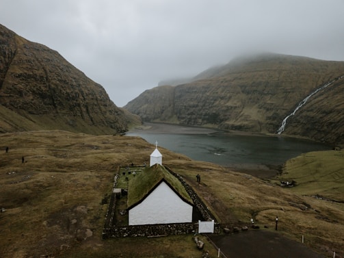 A small, white church with a grass roof is situated in a vast, open landscape characterized by large, rugged hills and a foggy sky. A waterfall cascades down one of the hills into a calm body of water, contributing to the serene and remote atmosphere. The terrain surrounding the church is grassy and uneven, with a few scattered rocks and pathways.