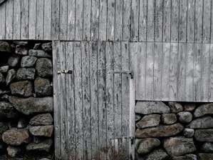 An old, weathered wooden door is set into a structure made of large, irregular stones. The planks of the door and nearby wooden siding show varied textures and grain patterns, suggesting age and exposure to the elements. The stones are rugged, with earthy tones and hints of moss, contributing to a rustic and historic appearance.