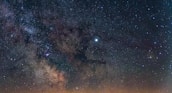 A vivid depiction of the Milky Way galaxy with countless stars scattered across the night sky. The galaxy's core is clearly visible, showing a bright, concentrated area surrounded by interstellar dust clouds. The image captures the vastness and beauty of the cosmos.