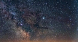 A vivid depiction of the Milky Way galaxy with countless stars scattered across the night sky. The galaxy's core is clearly visible, showing a bright, concentrated area surrounded by interstellar dust clouds. The image captures the vastness and beauty of the cosmos.