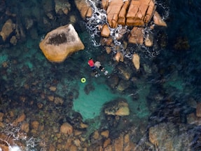 A person snorkeling in clear turquoise waters surrounded by large rocks. The top view emphasizes the rugged texture of the rocks beneath the water surface, providing a contrast between the calm sea and the vibrant red of the snorkeler's attire. A floating green ring is nearby, adding a pop of color to the natural scene.