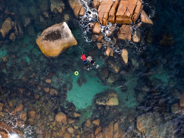 A person snorkeling in clear turquoise waters surrounded by large rocks. The top view emphasizes the rugged texture of the rocks beneath the water surface, providing a contrast between the calm sea and the vibrant red of the snorkeler's attire. A floating green ring is nearby, adding a pop of color to the natural scene.