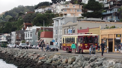 A coastal street scene features a vintage-style trolley bus with 'Big Bus San Francisco' signage. People are walking and crossing the street near parked cars. Buildings of various styles and colors, including a distinctive blue one, are lined up along the road. The background showcases hillside residential areas covered in lush greenery.