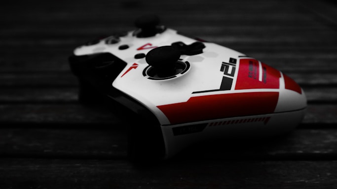 A video game controller with a sleek design, predominantly white with red and black accents. It sits on a dark wooden surface, with shadows enhancing its contours and details.
