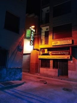 A dimly lit street corner at night, featuring a neon sign for 'Cine Arenas' attached to a building. Adjacent is a cafe with a yellow banner advertising breakfasts, tapas, and combined dishes. The street and buildings are bathed in a mix of blue and orange artificial lighting, creating a moody ambiance.