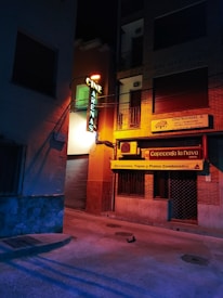 A dimly lit street corner at night, featuring a neon sign for 'Cine Arenas' attached to a building. Adjacent is a cafe with a yellow banner advertising breakfasts, tapas, and combined dishes. The street and buildings are bathed in a mix of blue and orange artificial lighting, creating a moody ambiance.
