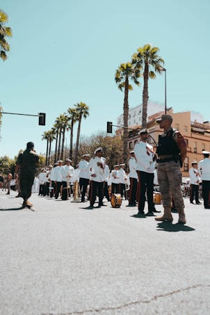 A marching band dressed in white uniforms is standing on a street lined with tall palm trees. The band members hold musical instruments, including brass instruments and drums. Two individuals in military-style attire are standing at the forefront, seemingly overseeing the event. The background features buildings and a clear, blue sky.