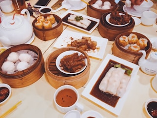 An assortment of dim sum dishes is arranged on a table. Wooden steamers hold items like buns and dumplings, while small plates display various dishes including chicken feet and rice noodles in sauce. A white teapot and cups filled with tea are also present, alongside dipping sauces.