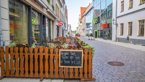 A cozy outdoor seating area is set up along a cobblestone street, enclosed by a wooden fence adorned with flower planters. A blackboard with text written in German invites passersby. The street is lined with buildings, including a modern glass-fronted one, and reflects a blend of old and new architecture.
