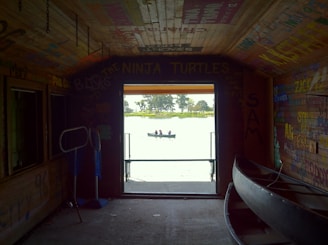 A rustic room with graffiti on wooden walls and ceiling, featuring phrases and names in various colors. Inside, a canoe is propped against the wall. Sunlight pours in through an open doorway, providing a view of a serene lake and a small boat with people in the distance.