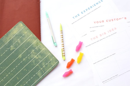 Two lined notebooks are placed on a white surface along with a few colorful gummy candies resembling fish shapes. Two pens with pastel colors are positioned diagonally. There are also printed sheets with headings such as 'The Experience', 'Your Customer', and 'The Big Idea'. The overall arrangement suggests a workspace setup.