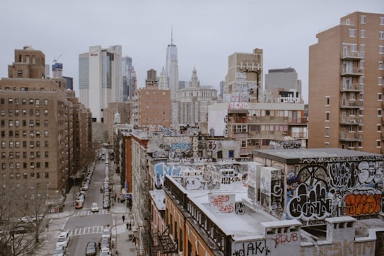 Urban landscape featuring numerous buildings with diverse architectural styles. Foreground buildings are heavily adorned with graffiti, giving a gritty, artistic vibe. Skyscrapers, including notable tall structures, rise in the background against an overcast sky. Streets below are lined with parked cars and pedestrians.