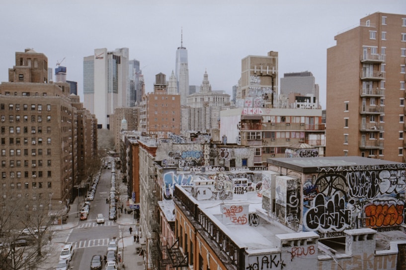 Urban landscape featuring numerous buildings with diverse architectural styles. Foreground buildings are heavily adorned with graffiti, giving a gritty, artistic vibe. Skyscrapers, including notable tall structures, rise in the background against an overcast sky. Streets below are lined with parked cars and pedestrians.