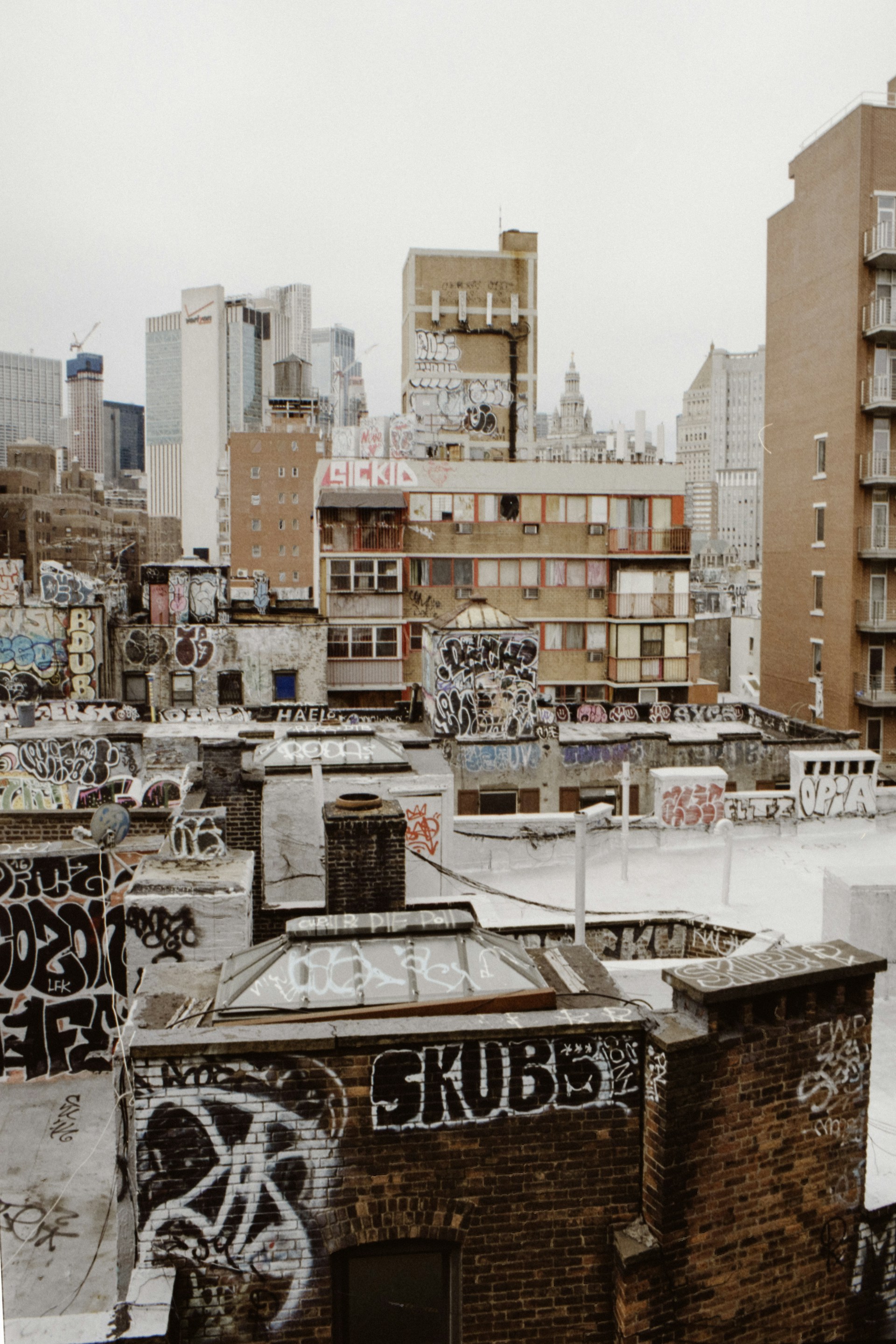 A dense urban area with multiple rooftops covered in graffiti. Older brick buildings contrast against a modern skyline in the background, which includes several high-rises. The foreground features a mix of flat and peaked rooftops adorned with various graffiti tags and art.