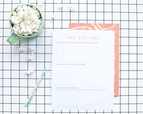A flat lay composition features a green mug filled with popcorn, placed on a white and black grid-patterned surface. Next to the mug is a worksheet titled 'The Big Idea' with sections for describing a business concept. A mint green pen lies nearby, and some scattered popcorn pieces are visible on the grid.
