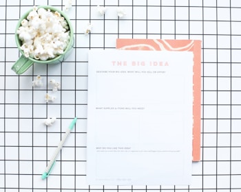 A flat lay composition features a green mug filled with popcorn, placed on a white and black grid-patterned surface. Next to the mug is a worksheet titled 'The Big Idea' with sections for describing a business concept. A mint green pen lies nearby, and some scattered popcorn pieces are visible on the grid.